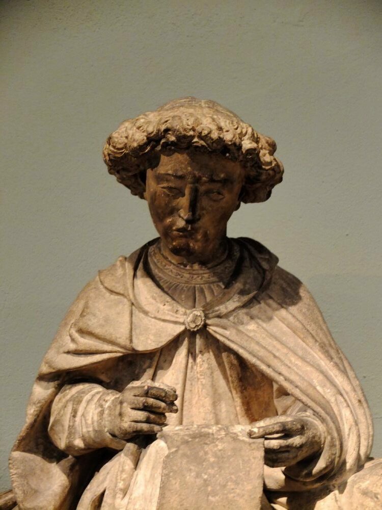 a medieval stone sculpture of a boy with curly hair, wearing a cloak and holding a piece of parchment