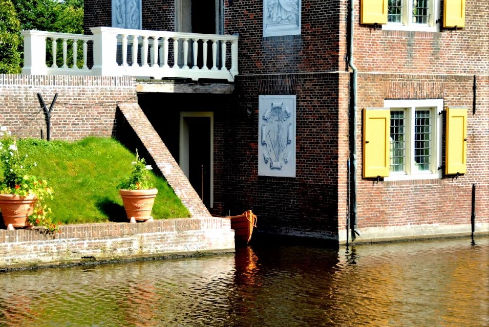 One corner of the Hofwijck mansion where it meets the water of the canal, reflecting in the water. Potted plants sit on a grassy bank nearby