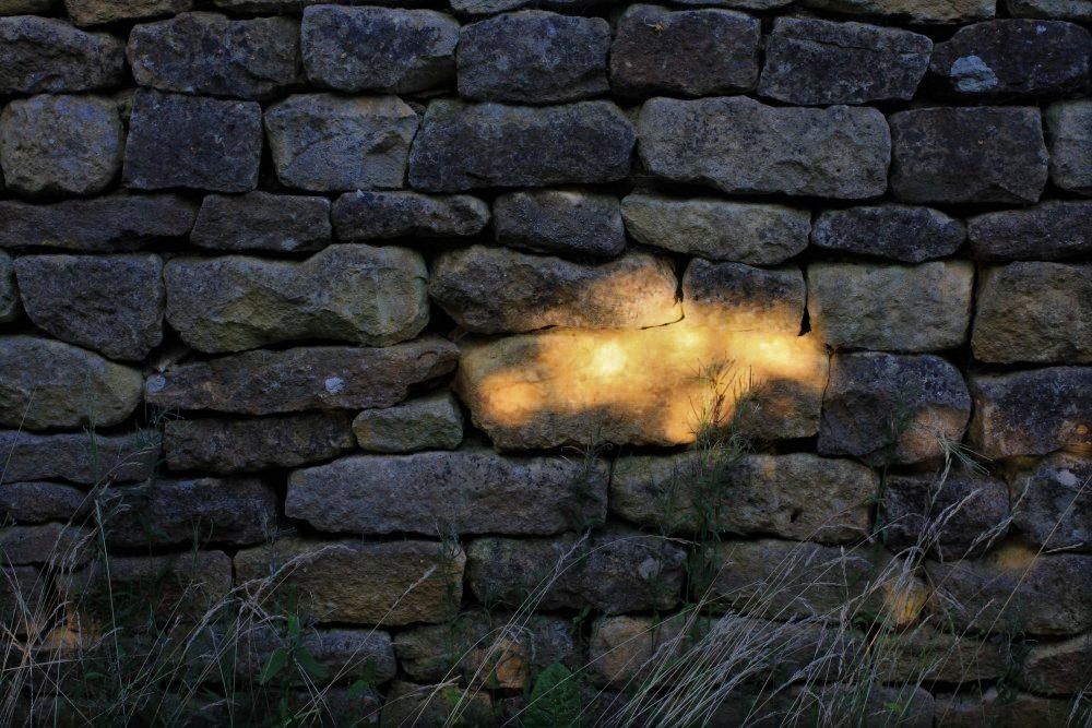 a stone wall cast in deep shadow, illuminated at the center by one small golden sun spot