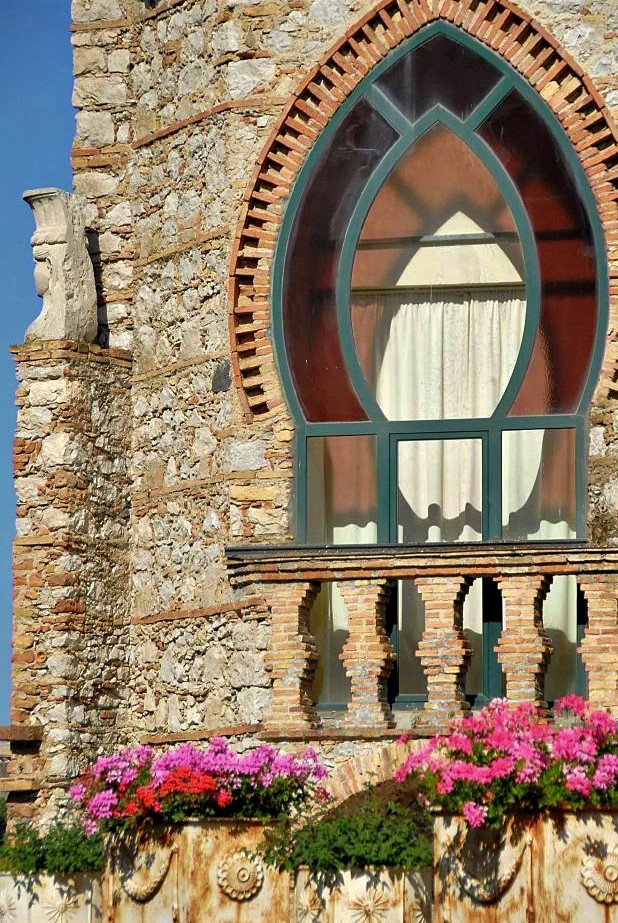 A stone house with a large ornate window in the shape of a pointed oval. Pink flowers sit in pots at the base of the house