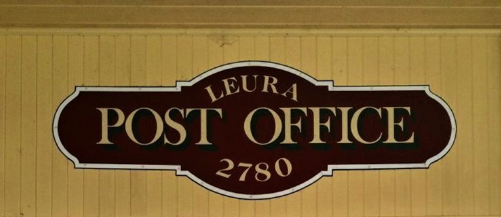A dark-brown antique sign that reads 'Leura Post Office 2780'. It hangs on a pale mustard-yellow wall.