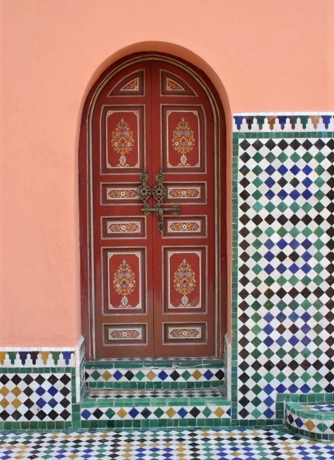 A brown wooden door with a curved top, surrounded by a peach-painted terra-cotta wall with intricately patterned tiles