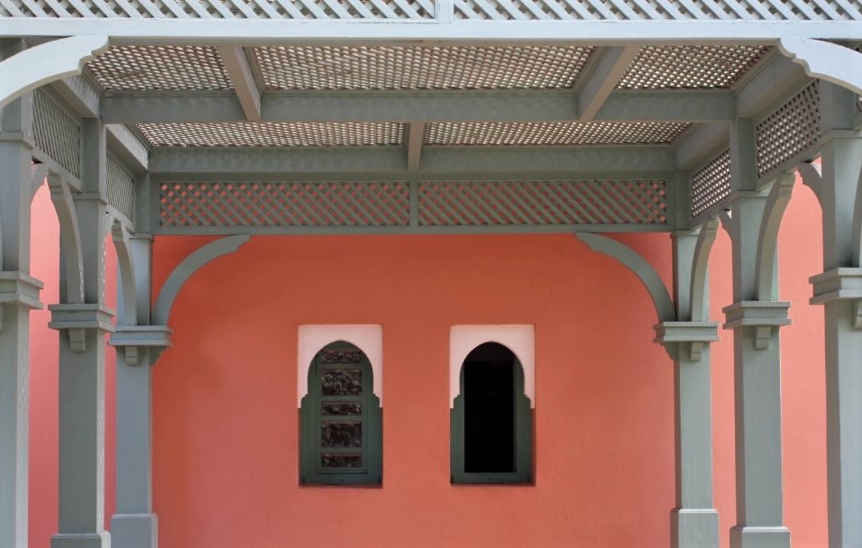 A peach-coloured terra-cotta wall with two bud-shaped windows, above which is a blue pergola