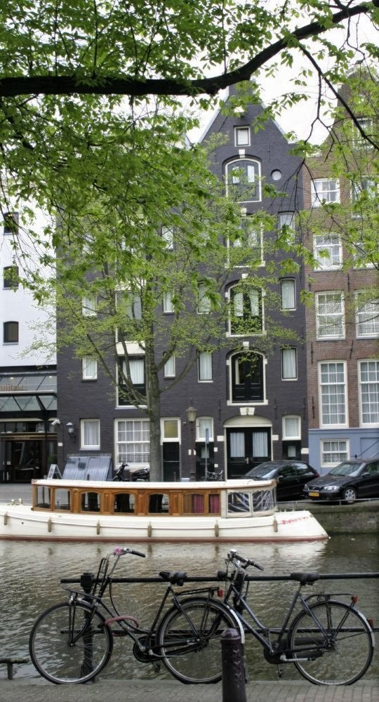 A row of bicycles lean against a metal railing in the foreground. Behind them a white boat floats in a canal in front of tall narrow buildings