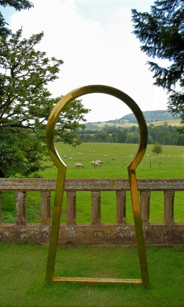 A large metal sculpture of a keyhole, located in an English garden. In the distance, a flock of sheep graze in a green meadow
