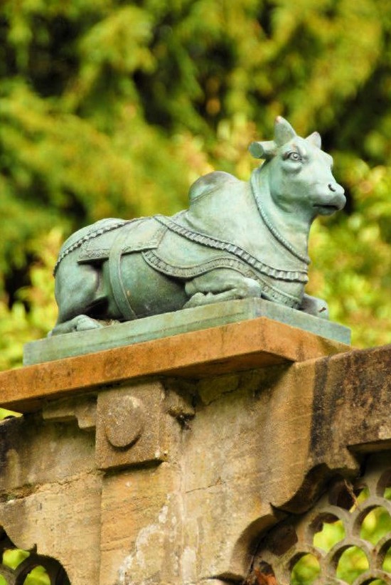 A copper statue of a bull sitting on a stone pedestal in a garden