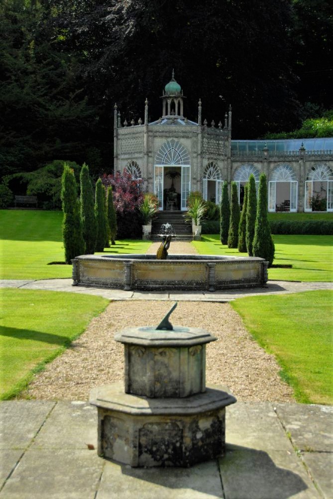 A sundial in a manicured garden the foreground with part of an ornate stone mansion visible in the background