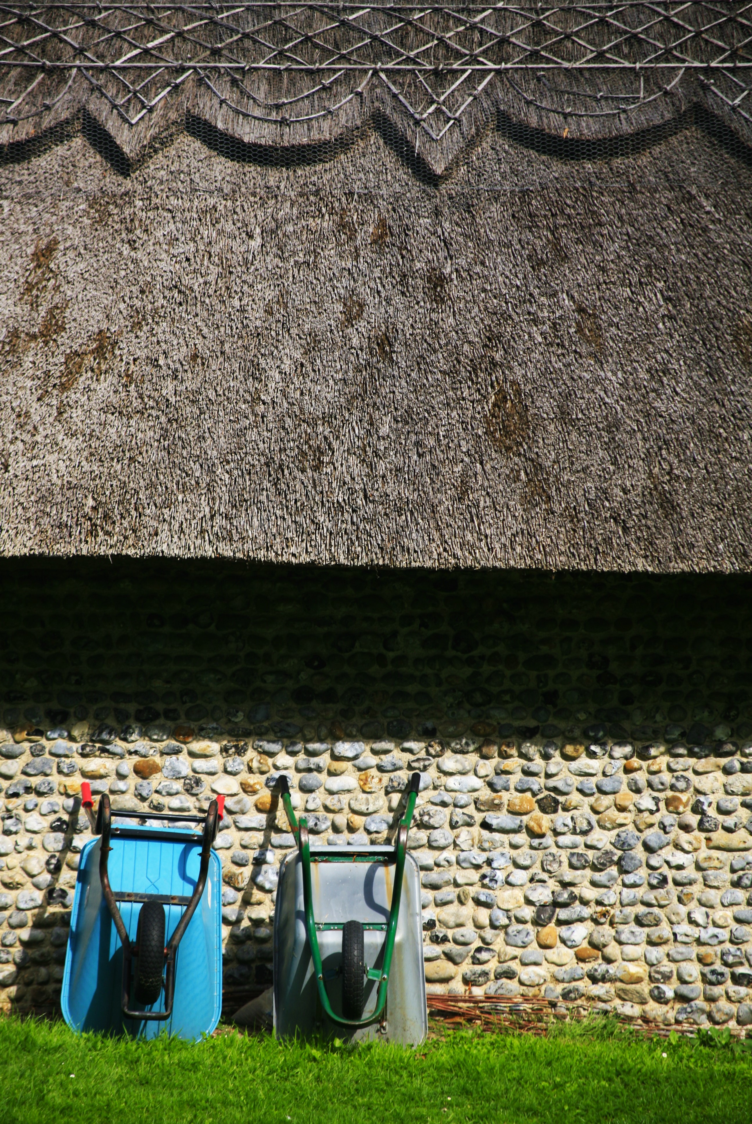 Two wheelbarrows leaning against the wall of a stone cottage with a thatched roof
