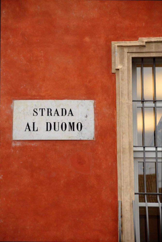 A dark salmon-coloured wall of a building with a plaque that reads, Strada al Duomo