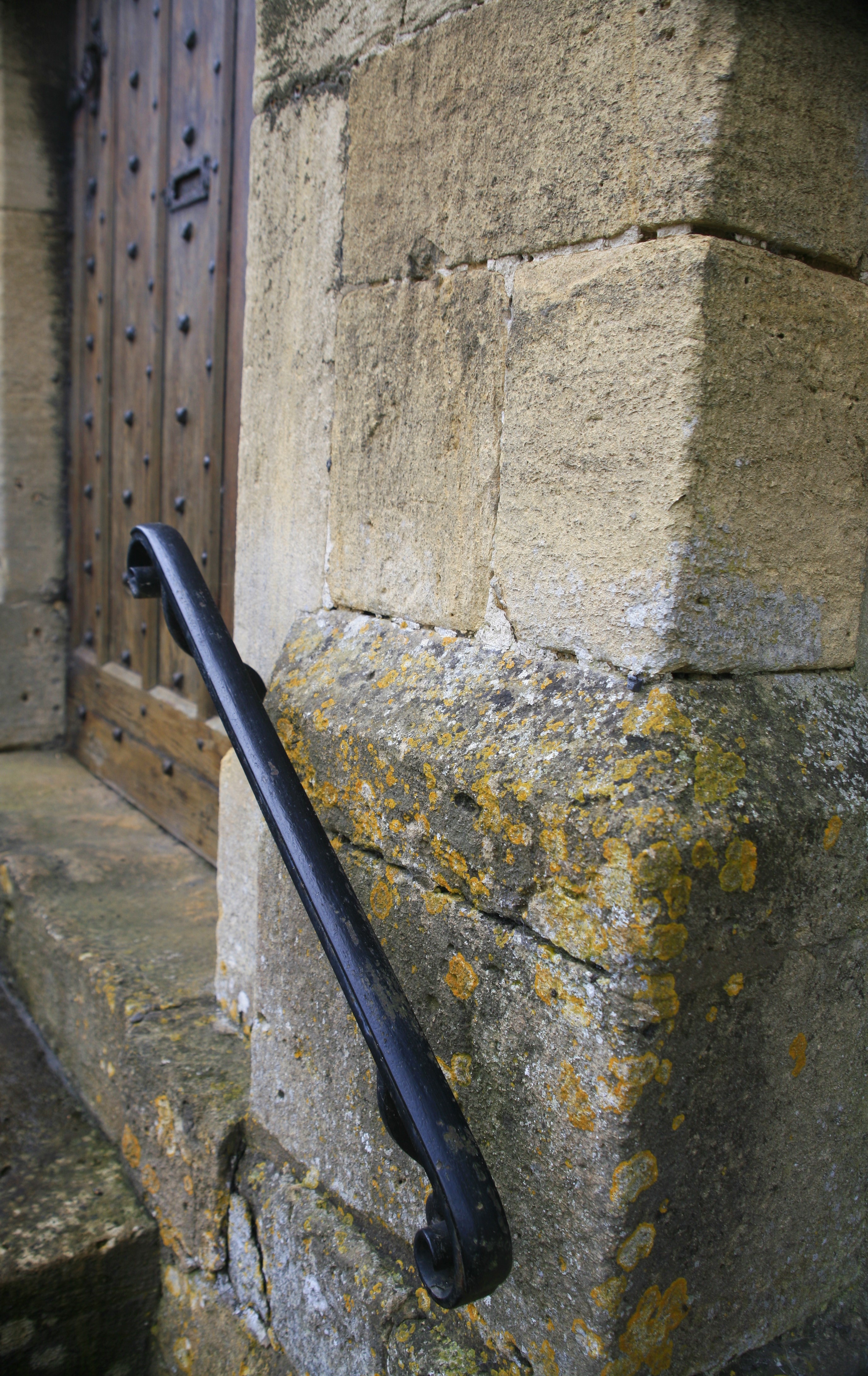 A metal railing fixed in a stone wall leading up to an old wooden door seen from a sharp angle