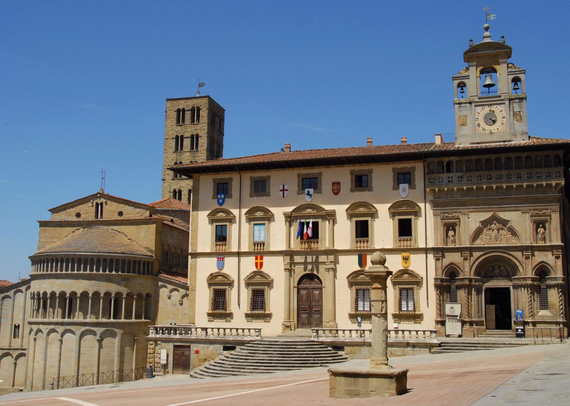 A village square featuring a beige stone building with many windows and various coats of arms decorating it