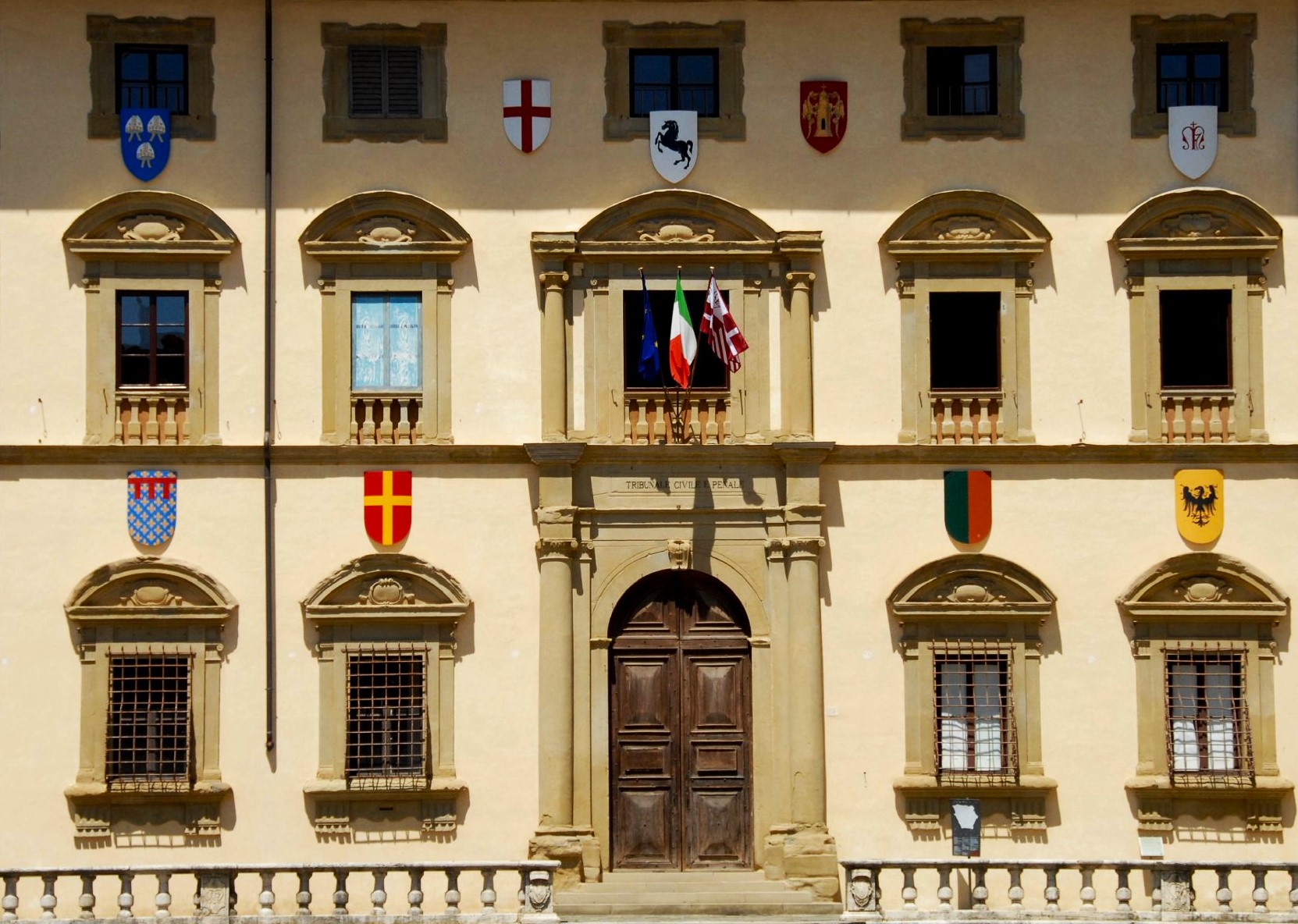 The facade of a beige stone building with many windows and various coats of arms decorating it